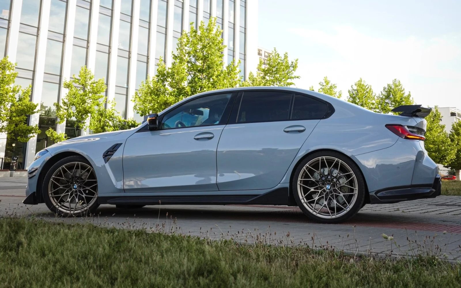 Šedé BMW M3 Competition z boku se zadním křídlem a bronzovými koly