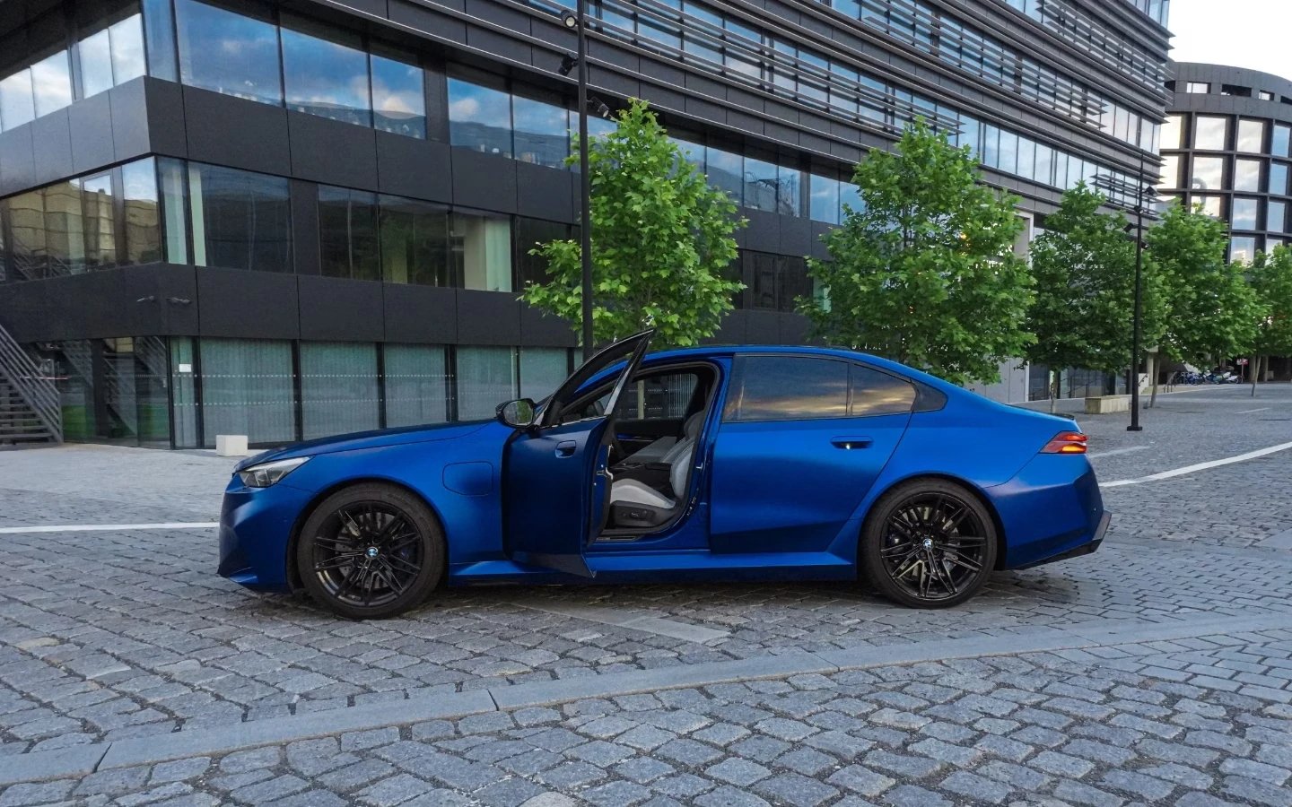 Modré BMW M5 z boku s otevřenými dveřmi před moderní prosklenou budovou